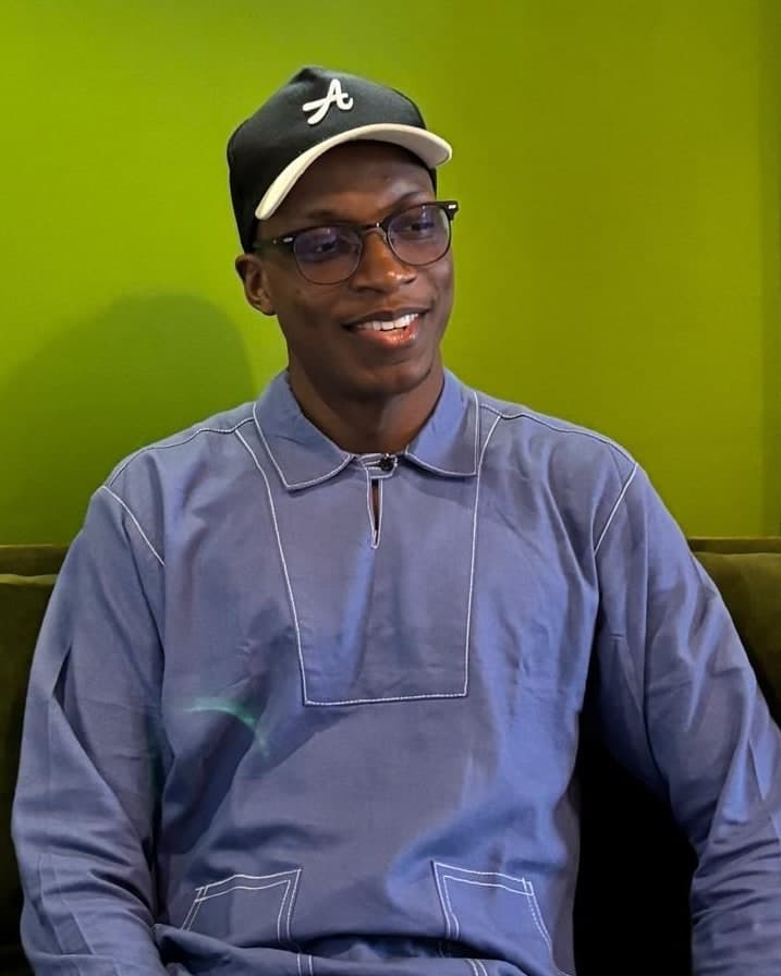 Akin smiling, in a blue shirt, glasses and a black cap, against a green wall.